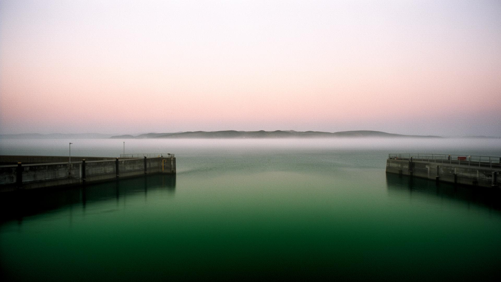 A quiet coastal harbor at dawn with mist over still water.